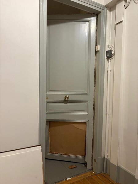 Porte intérieure grise avec panneau inférieur cassé comblé par un carton provisoire, chambranle endommagé